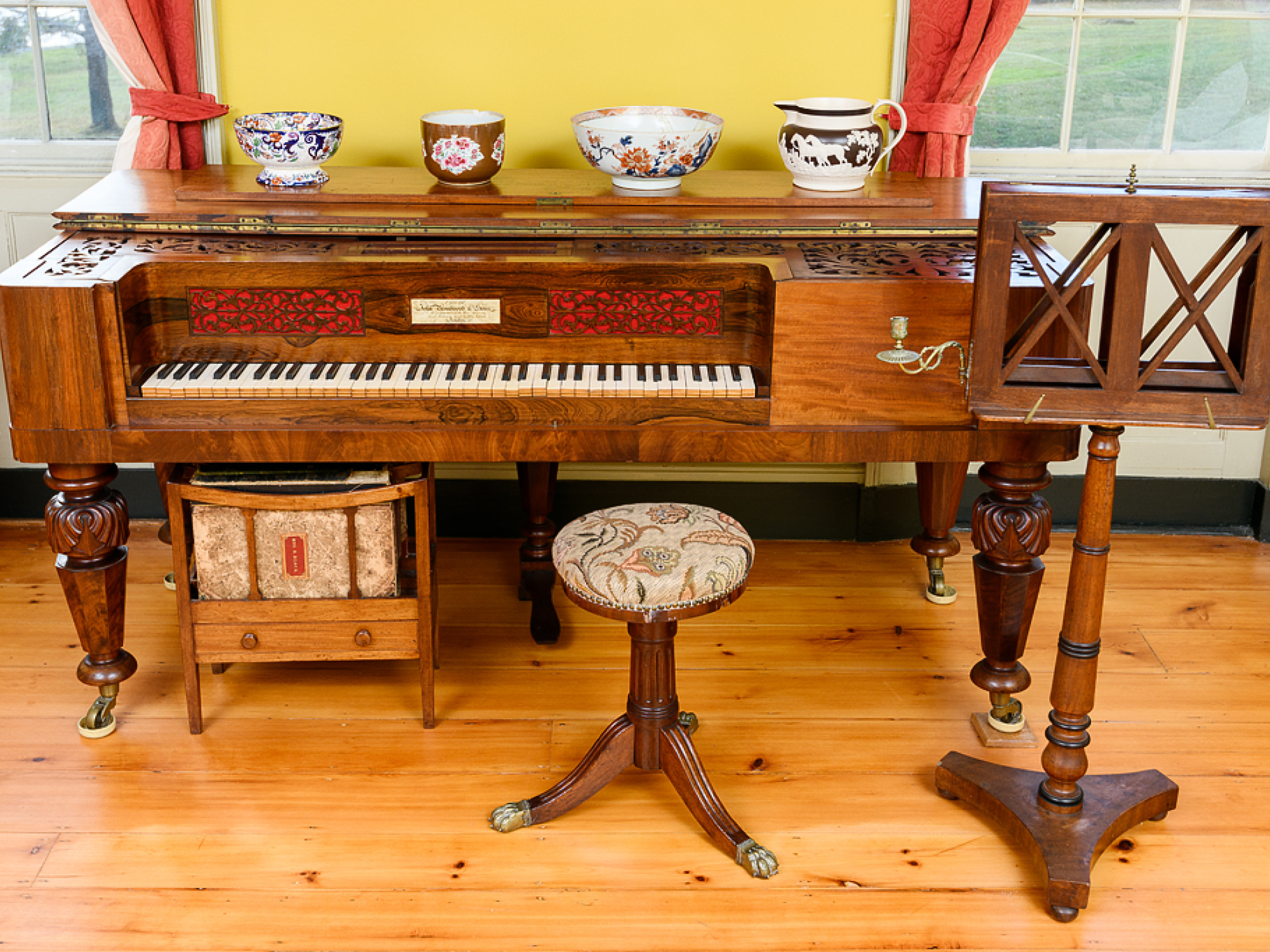 A small, ornate piano with decorative wood inlay and a matching stool, positioned in a room with colorful drapes and a wooden music stand.