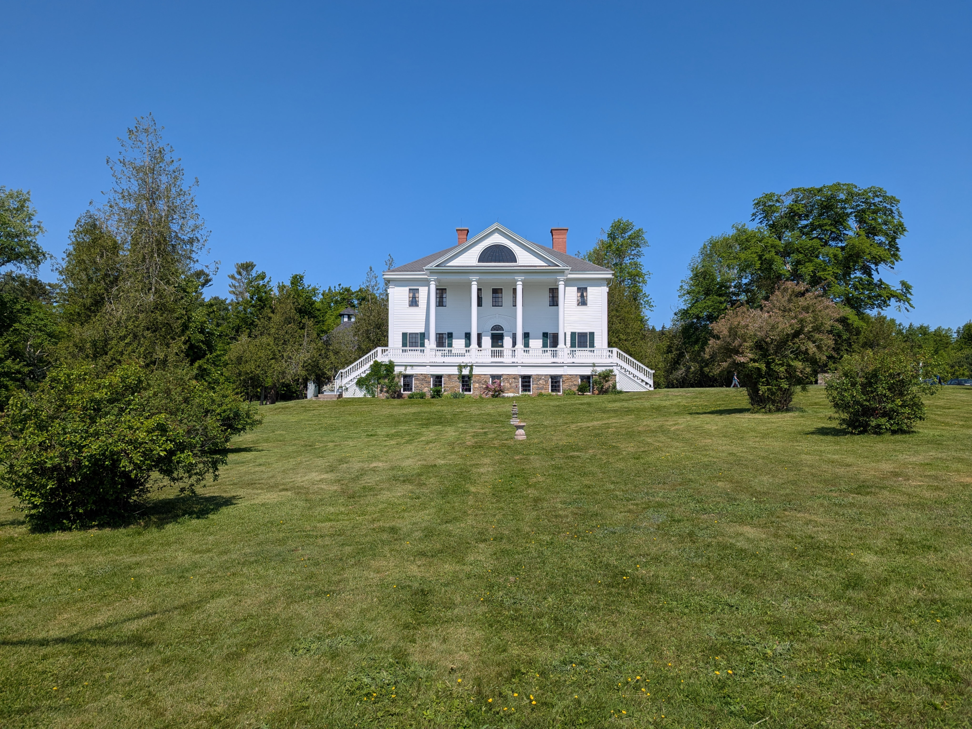 Uniacke Estate a grand white house with a classical design, including a columned entrance and a long veranda. The house is surrounded by a well-maintained lawn and mature trees.