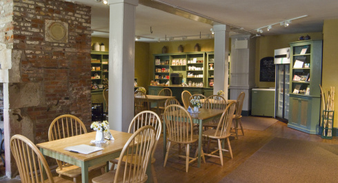 A bright and airy tea room with wooden tables and chairs, and shelves stocked with items.