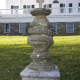 A stone monument with a cross on top, standing in front of a large white house.