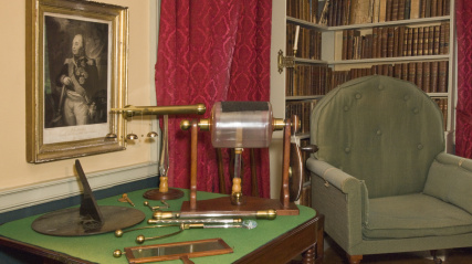 library featuring a tall bookcase, several paintings, and a table with a telescope-like device.