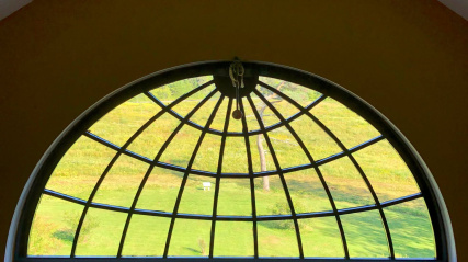 A large, semicircular fanlight window with a black grid pattern looks out onto a vibrant green field and rolling hills from inside a dimly lit room with wooden floors.
