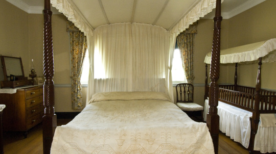 bedroom featuring a large four-poster canopy bed with an intricately patterned, light-colored quilt, accompanied by a wooden baby crib.
