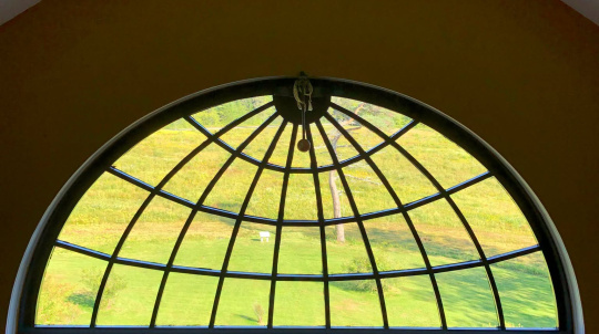 A large, semicircular fanlight window with a black grid pattern looks out onto a vibrant green field and rolling hills from inside a dimly lit room with wooden floors.